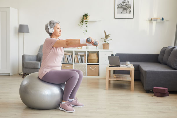 An older woman works out on a silver exercise ball while holding a small weight.