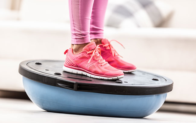 A close shot of a woman's feet in sneakers, standing on a balance ball doing vestibular physiotherapy.
